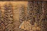 Carved wooden relief depicting a dense forest scene with tall, detailed pine trees surrounding a calm body of water. The intricate designs highlight the texture of the bark and the leaves, giving a sense of depth and natural beauty. Rocks are scattered along the edge, enhancing the rustic, wilderness setting.