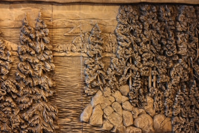Carved wooden relief depicting a dense forest scene with tall, detailed pine trees surrounding a calm body of water. The intricate designs highlight the texture of the bark and the leaves, giving a sense of depth and natural beauty. Rocks are scattered along the edge, enhancing the rustic, wilderness setting.