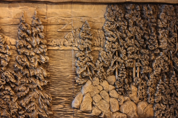 Carved wooden relief depicting a dense forest scene with tall, detailed pine trees surrounding a calm body of water. The intricate designs highlight the texture of the bark and the leaves, giving a sense of depth and natural beauty. Rocks are scattered along the edge, enhancing the rustic, wilderness setting.