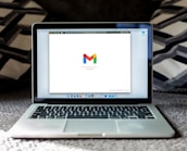 An open laptop on a carpeted surface displays the Gmail loading screen. The background is blurred, with patterned pillows or cushions adding a cozy setting.