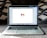 An open laptop on a carpeted surface displays the Gmail loading screen. The background is blurred, with patterned pillows or cushions adding a cozy setting.