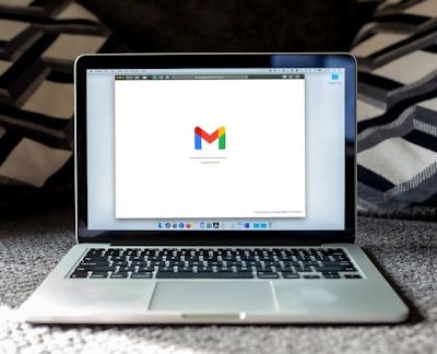 An open laptop on a carpeted surface displays the Gmail loading screen. The background is blurred, with patterned pillows or cushions adding a cozy setting.