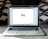 An open laptop on a carpeted surface displays the Gmail loading screen. The background is blurred, with patterned pillows or cushions adding a cozy setting.