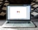 An open laptop on a carpeted surface displays the Gmail loading screen. The background is blurred, with patterned pillows or cushions adding a cozy setting.