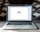 An open laptop on a carpeted surface displays the Gmail loading screen. The background is blurred, with patterned pillows or cushions adding a cozy setting.