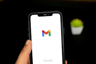 A hand holding a smartphone displaying the Gmail logo on its screen, against a dark background with a small green plant blurred in the background.