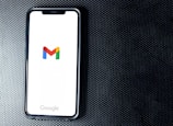 A smartphone with a white screen displays the Gmail logo, positioned on a textured dark background, with the Google branding at the bottom of the screen.