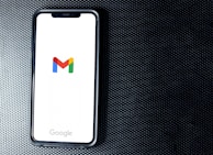 A smartphone with a white screen displays the Gmail logo, positioned on a textured dark background, with the Google branding at the bottom of the screen.