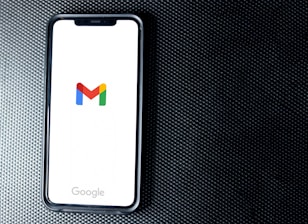 A smartphone with a white screen displays the Gmail logo, positioned on a textured dark background, with the Google branding at the bottom of the screen.