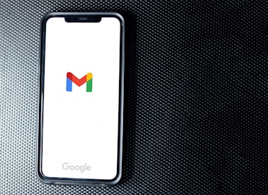 A smartphone with a white screen displays the Gmail logo, positioned on a textured dark background, with the Google branding at the bottom of the screen.