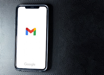 A smartphone with a white screen displays the Gmail logo, positioned on a textured dark background, with the Google branding at the bottom of the screen.