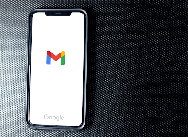 A smartphone with a white screen displays the Gmail logo, positioned on a textured dark background, with the Google branding at the bottom of the screen.