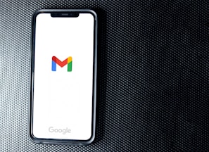 A smartphone with a white screen displays the Gmail logo, positioned on a textured dark background, with the Google branding at the bottom of the screen.