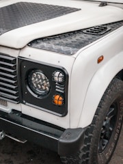 A front view of a rugged off-road vehicle, featuring a white body with black textured panels and trim. The image highlights the right headlight, indicator lights, and portion of the wheel with large, knobby tires. The vehicle has a utilitarian design with strong lines and a durable construction.