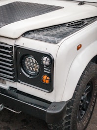 A front view of a rugged off-road vehicle, featuring a white body with black textured panels and trim. The image highlights the right headlight, indicator lights, and portion of the wheel with large, knobby tires. The vehicle has a utilitarian design with strong lines and a durable construction.