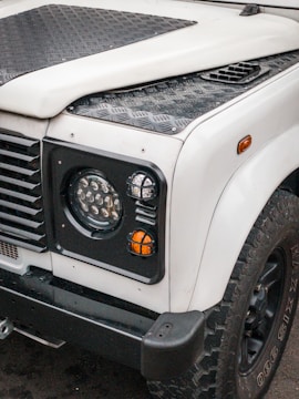 A front view of a rugged off-road vehicle, featuring a white body with black textured panels and trim. The image highlights the right headlight, indicator lights, and portion of the wheel with large, knobby tires. The vehicle has a utilitarian design with strong lines and a durable construction.
