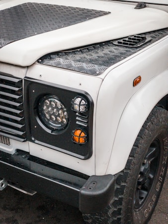 A front view of a rugged off-road vehicle, featuring a white body with black textured panels and trim. The image highlights the right headlight, indicator lights, and portion of the wheel with large, knobby tires. The vehicle has a utilitarian design with strong lines and a durable construction.