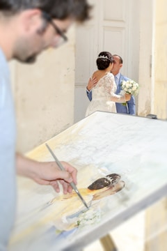 A couple is embracing and kissing in a bright, elegantly decorated space. The bride is wearing a white, lace wedding dress and holding a bouquet of flowers. In the foreground, there's an artist painting a portrait of the couple on a canvas, capturing the romantic moment.