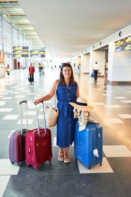 A woman is standing in an airport terminal with three large suitcases, two red and one blue, along with a woven bag. She is wearing a denim dress and sandals, with a sun hat placed on the handle of one suitcase. The terminal is spacious and bright, featuring tiled floors and overhead flight information displays.