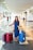 A woman is standing in an airport terminal with three large suitcases, two red and one blue, along with a woven bag. She is wearing a denim dress and sandals, with a sun hat placed on the handle of one suitcase. The terminal is spacious and bright, featuring tiled floors and overhead flight information displays.