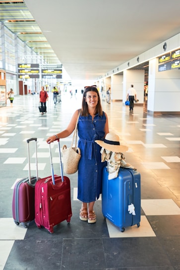 A woman is standing in an airport terminal with three large suitcases, two red and one blue, along with a woven bag. She is wearing a denim dress and sandals, with a sun hat placed on the handle of one suitcase. The terminal is spacious and bright, featuring tiled floors and overhead flight information displays.