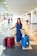 A woman is standing in an airport terminal with three large suitcases, two red and one blue, along with a woven bag. She is wearing a denim dress and sandals, with a sun hat placed on the handle of one suitcase. The terminal is spacious and bright, featuring tiled floors and overhead flight information displays.