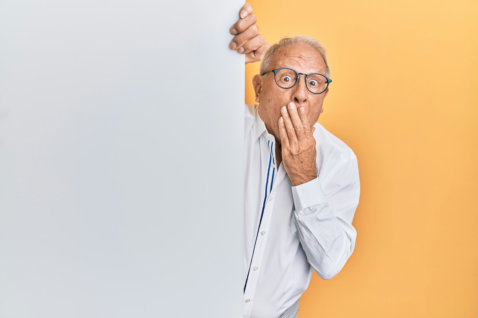 Senior caucasian man holding blank empty banner covering mouth with hand, shocked and afraid for mistake. surprised expression 