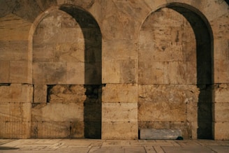 A stone wall with two large, rounded arches, constructed from unevenly colored bricks. The texture of the stone is rugged and weathered, giving it an aged appearance. There is a stone ledge or seat at the base of the right arch, partially in shadow.