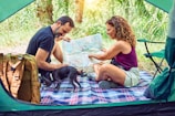 A couple sits inside a tent looking at a map together, surrounded by nature. They are seated on a checkered blanket with a small black dog. A hiking backpack is visible, suggesting an outdoor adventure or camping trip.