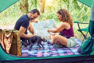 A couple sits inside a tent looking at a map together, surrounded by nature. They are seated on a checkered blanket with a small black dog. A hiking backpack is visible, suggesting an outdoor adventure or camping trip.