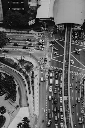 Aerial view of a busy urban intersection with numerous cars and motorcycles navigating through marked lanes. The area is surrounded by high-rise buildings and features a large transportation infrastructure, possibly a train or bus station, above the roadways. Sidewalks and landscaped areas are visible, with some pedestrians walking.