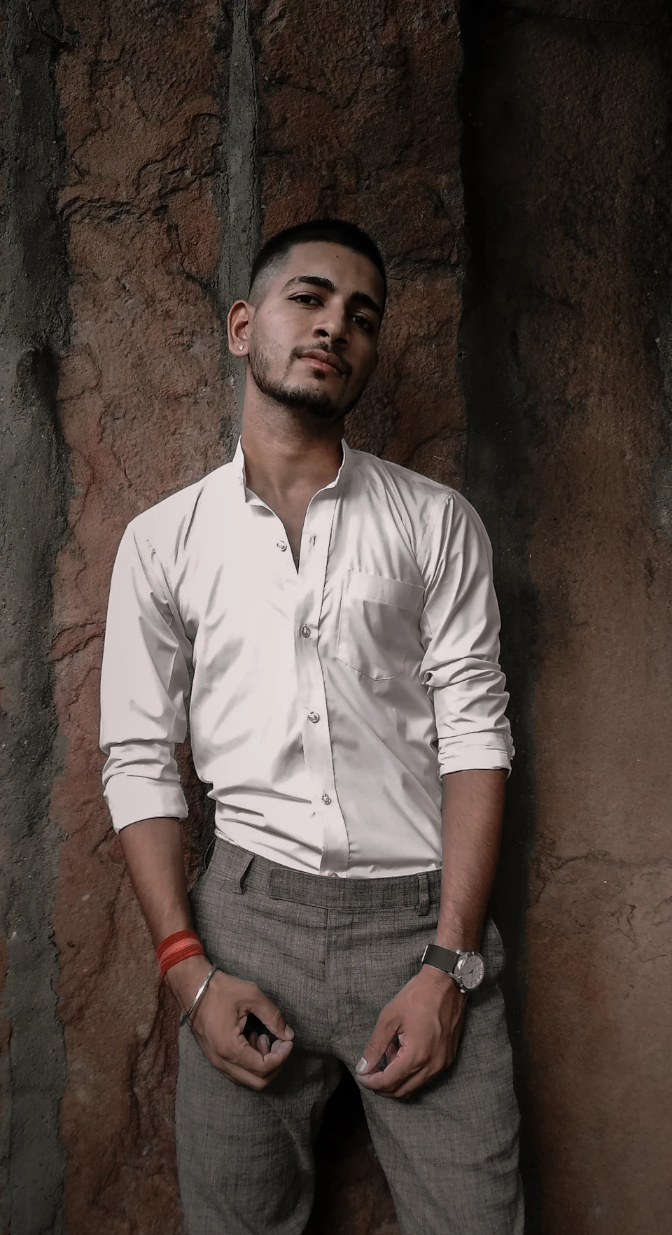 The image shows a young man standing against a stone wall, dressed in a white shirt and gray pants. He is looking directly at the camera with an expression of concentration or contemplation. The stone wall behind him has a rough texture, adding to the overall ruggedness of the scene.