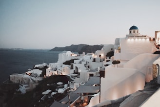 A picturesque coastal town with whitewashed buildings features prominently against a backdrop of calm, deep blue sea. The iconic blue domes and narrow winding paths are hallmark features of a Mediterranean landscape. The evening ambiance is set by warm lights illuminating the structures, casting soft glows that highlight the architecture and surrounding cliffs.