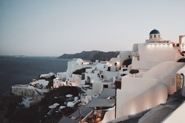 A picturesque coastal town with whitewashed buildings features prominently against a backdrop of calm, deep blue sea. The iconic blue domes and narrow winding paths are hallmark features of a Mediterranean landscape. The evening ambiance is set by warm lights illuminating the structures, casting soft glows that highlight the architecture and surrounding cliffs.