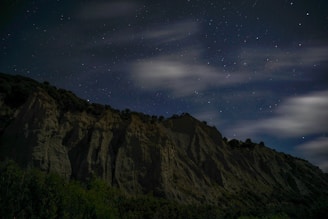 A rugged mountain ridge under a starry night sky, with wispy clouds partially obscuring the stars. The silhouette of the rocky cliff is highlighted by the faint ambient light, and dense vegetation is visible at the base of the cliffs.
