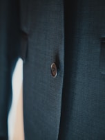 A close-up view of a dark-colored, textured fabric with a single, dark button visible. The fabric appears to be part of a garment, likely a suit jacket or coat.