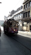 A vintage tram moves along the cobblestone streets of a European city, surrounded by historic buildings with ornate balconies. Pedestrians walk nearby, and the tram displays an advertisement. The street is illuminated by daylight, casting shadows.