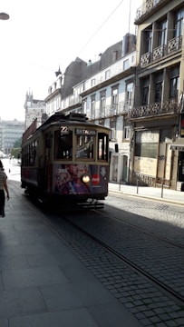 A vintage tram moves along the cobblestone streets of a European city, surrounded by historic buildings with ornate balconies. Pedestrians walk nearby, and the tram displays an advertisement. The street is illuminated by daylight, casting shadows.