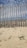 A wooden fence placed on sandy ground with sparse greenery scattered across the sand. The sky is blue with scattered clouds, creating a calm and serene atmosphere.