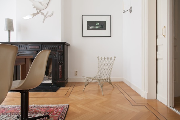 A minimalist interior space featuring a black wooden fireplace with intricate designs against a white wall. A unique metallic chair with a wireframe design is placed on the wooden parquet floor. A rectangular photograph in a black frame hangs above the chair. A modern, sculptural wall lamp shaped like a bird is mounted above the fireplace. Part of a wooden table with beige chairs and a red patterned rug is visible on the left.
