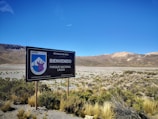 A landscape featuring a large welcome sign for Parque Nacional Lauca in Chile, situated in an arid desert environment. Sparse vegetation covers the foreground, while mountain ranges with varying shades of brown and red are visible in the background under a clear blue sky.