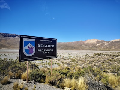 A landscape featuring a large welcome sign for Parque Nacional Lauca in Chile, situated in an arid desert environment. Sparse vegetation covers the foreground, while mountain ranges with varying shades of brown and red are visible in the background under a clear blue sky.
