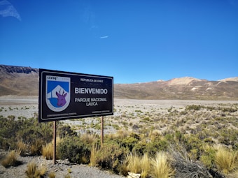 A landscape featuring a large welcome sign for Parque Nacional Lauca in Chile, situated in an arid desert environment. Sparse vegetation covers the foreground, while mountain ranges with varying shades of brown and red are visible in the background under a clear blue sky.