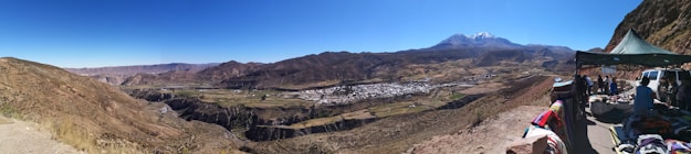 A panoramic view of a rugged mountain landscape with a clear blue sky. In the foreground, there is a market stall under a green tent where several people are gathered. To the left, there are expansive fields and a small town nestled in a valley. A snow-capped mountain peak rises in the distance.