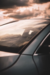 A sleek car window reflects a dramatic sky filled with moody clouds. The focus is on the window and a part of the car's body, showcasing smooth lines and a shiny surface.