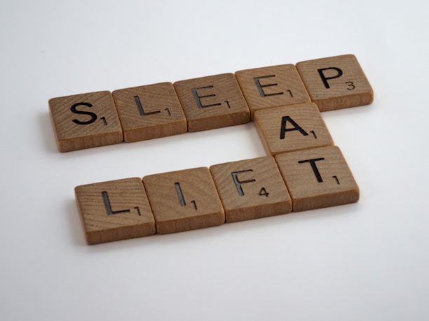 Wooden Scrabble tiles spell out the words SLEEP, EAT, and LIFT on a white background.