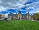 A grand historic estate with intricate architectural features and a large, manicured lawn in the foreground. The sky is partly cloudy with patches of blue peeking through. People are scattered across the lawn, and a tour bus is parked to the side.