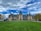 A grand historic estate with intricate architectural features and a large, manicured lawn in the foreground. The sky is partly cloudy with patches of blue peeking through. People are scattered across the lawn, and a tour bus is parked to the side.