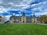 A grand historic estate with intricate architectural features and a large, manicured lawn in the foreground. The sky is partly cloudy with patches of blue peeking through. People are scattered across the lawn, and a tour bus is parked to the side.