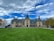 A grand historic estate with intricate architectural features and a large, manicured lawn in the foreground. The sky is partly cloudy with patches of blue peeking through. People are scattered across the lawn, and a tour bus is parked to the side.